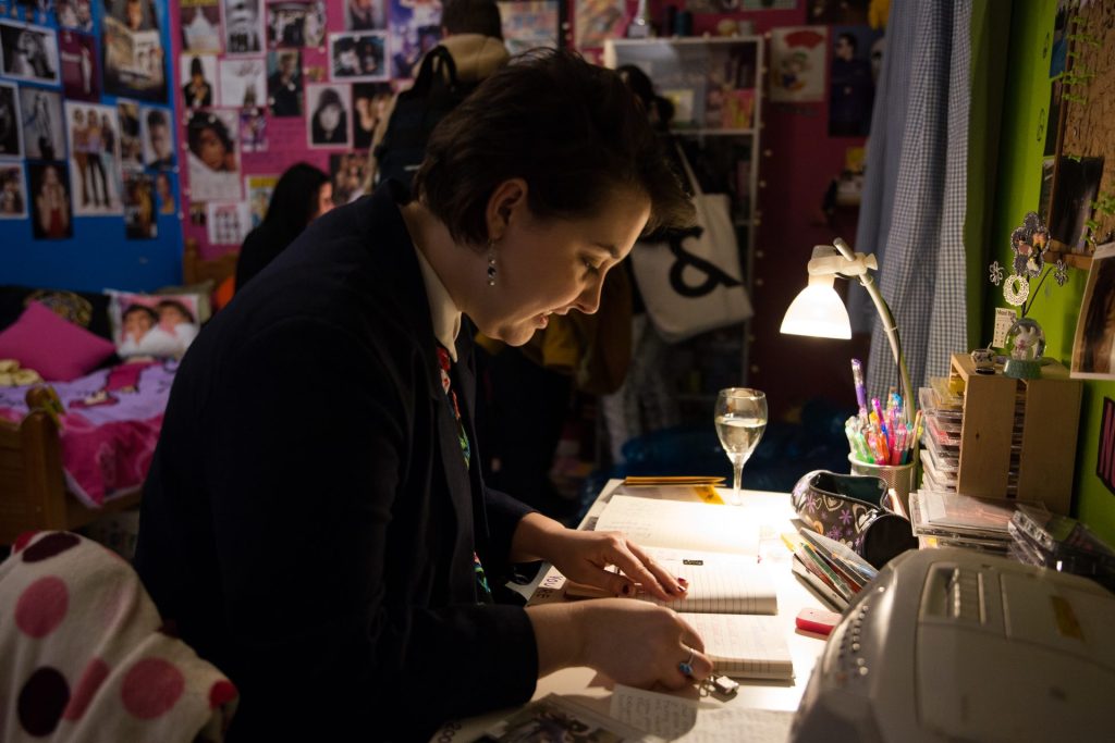 A person sits at a cluttered desk under a lamp, focused on writing in a notebook. The room is filled with posters and personal items, creating a cozy, creative atmosphere.