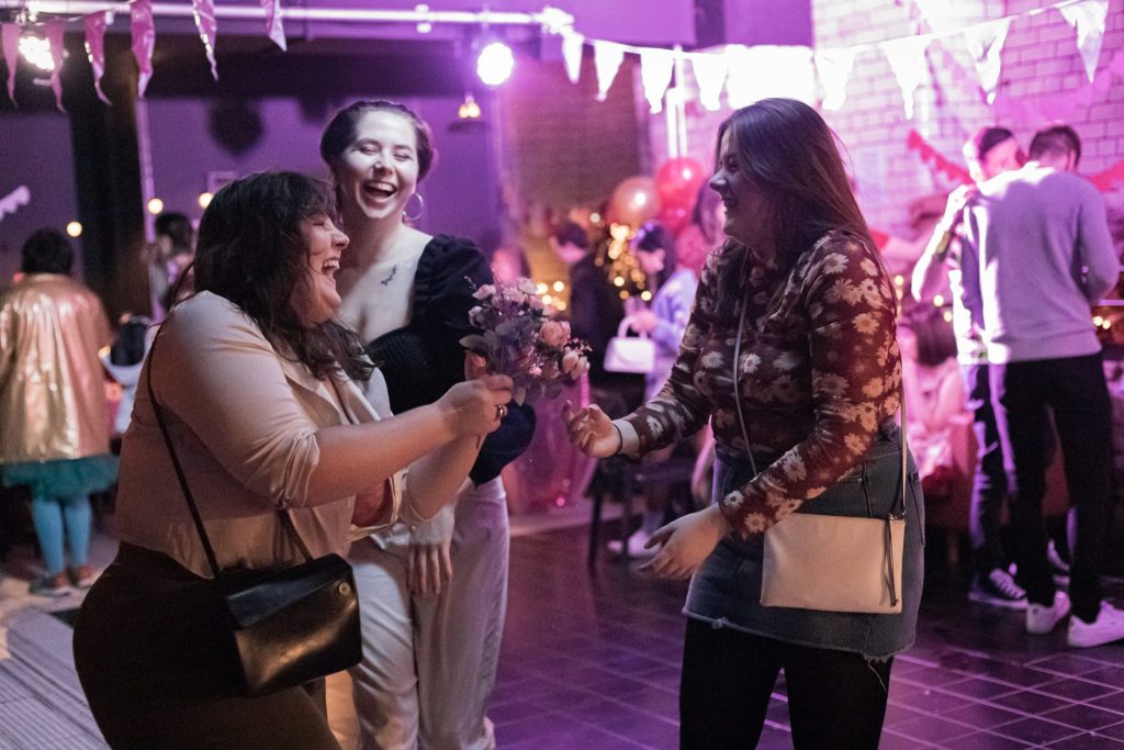 Three people joyfully dance and laugh in a festively decorated room with balloons and streamers. One is holding a bouquet. The scene is lively with others socializing in the background.