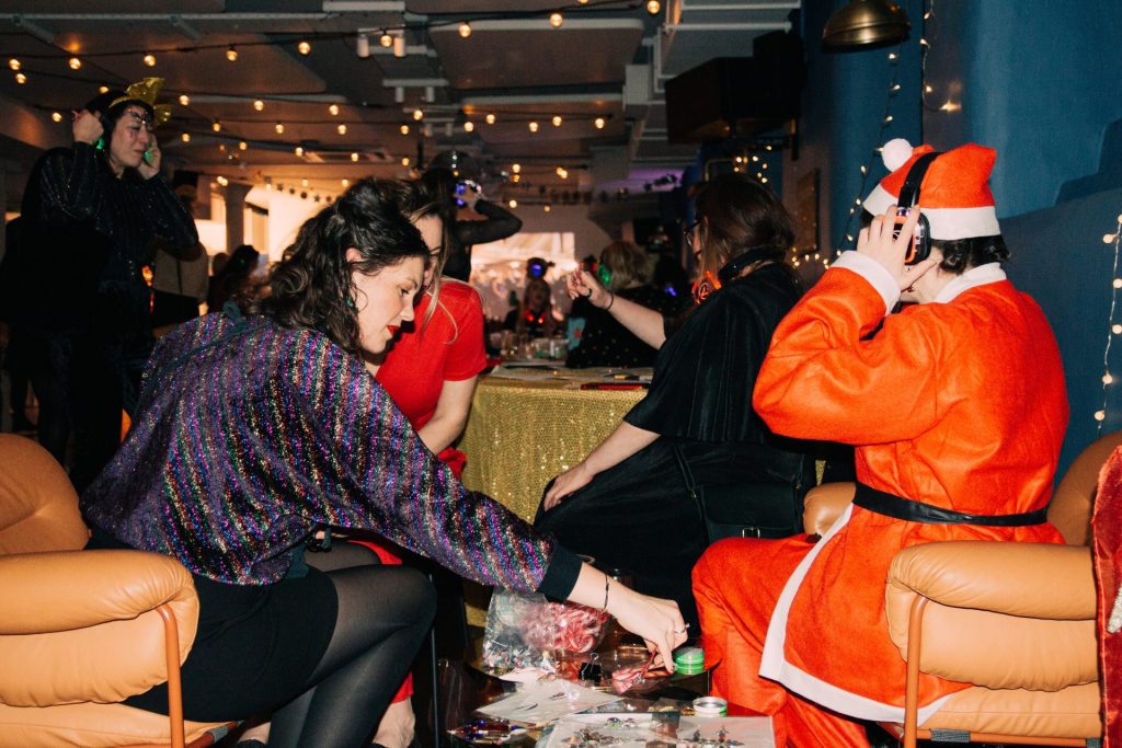 People enjoy a festive party with a holiday theme. One person wears a Santa Claus costume while others are seated and interacting in a room decorated with string lights. A table with various items is in the foreground.