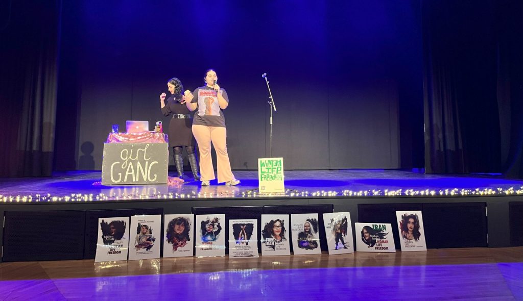 Two people stand on a stage under blue lighting. One is at a laptop on a decorated table, while the other holds a microphone. In front are posters with various images and text. A sign on the table reads "Girl Gang.