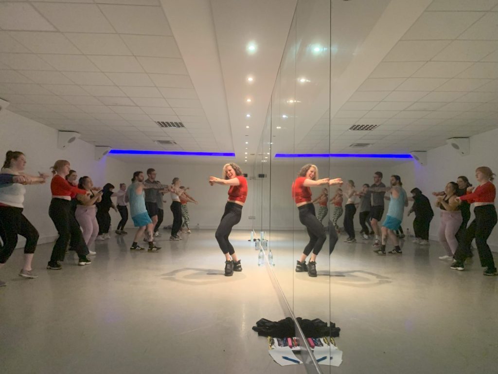 group of people dancing in front of a woman leading the class all reflected in a mirror wall behind the woman