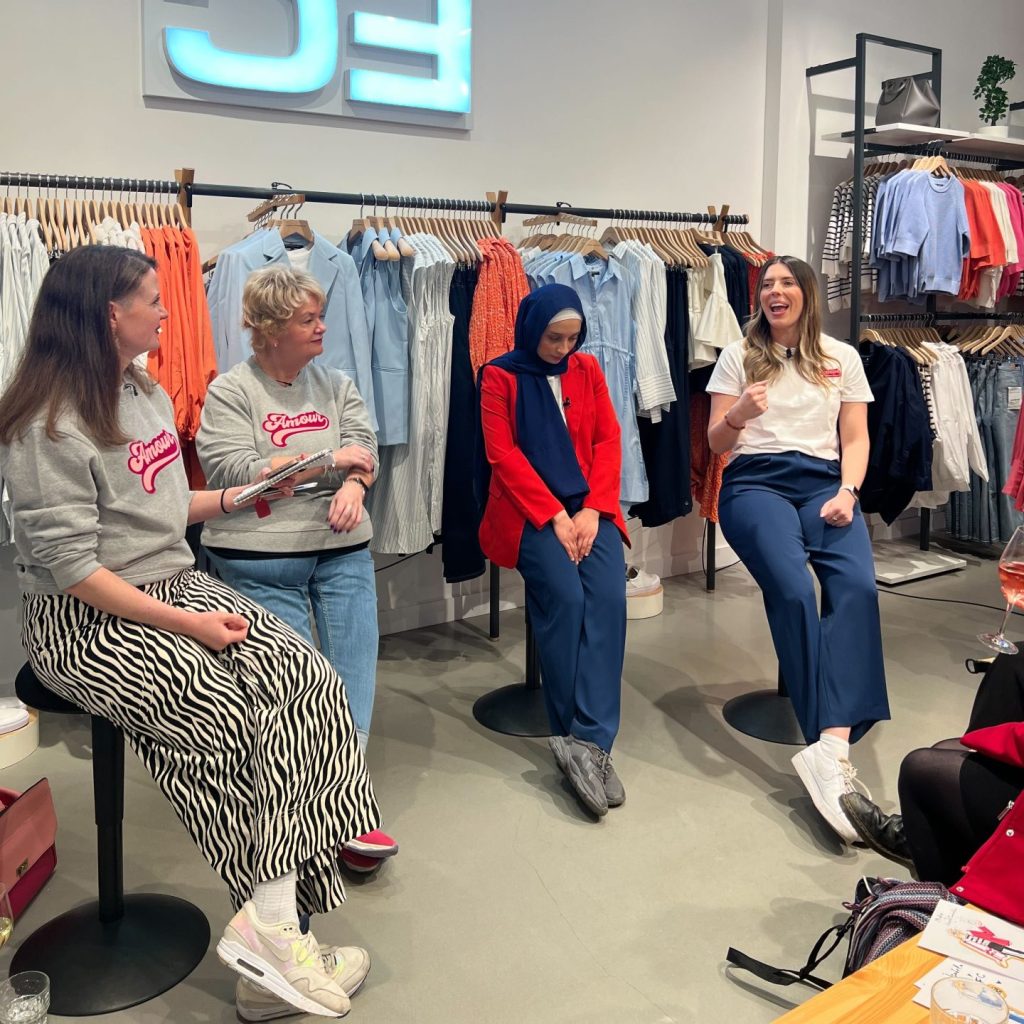 Four women sit on stools in a clothing store, engaged in conversation. Behind them, racks of clothes and a bright logo on the wall are visible. The atmosphere is casual and friendly.