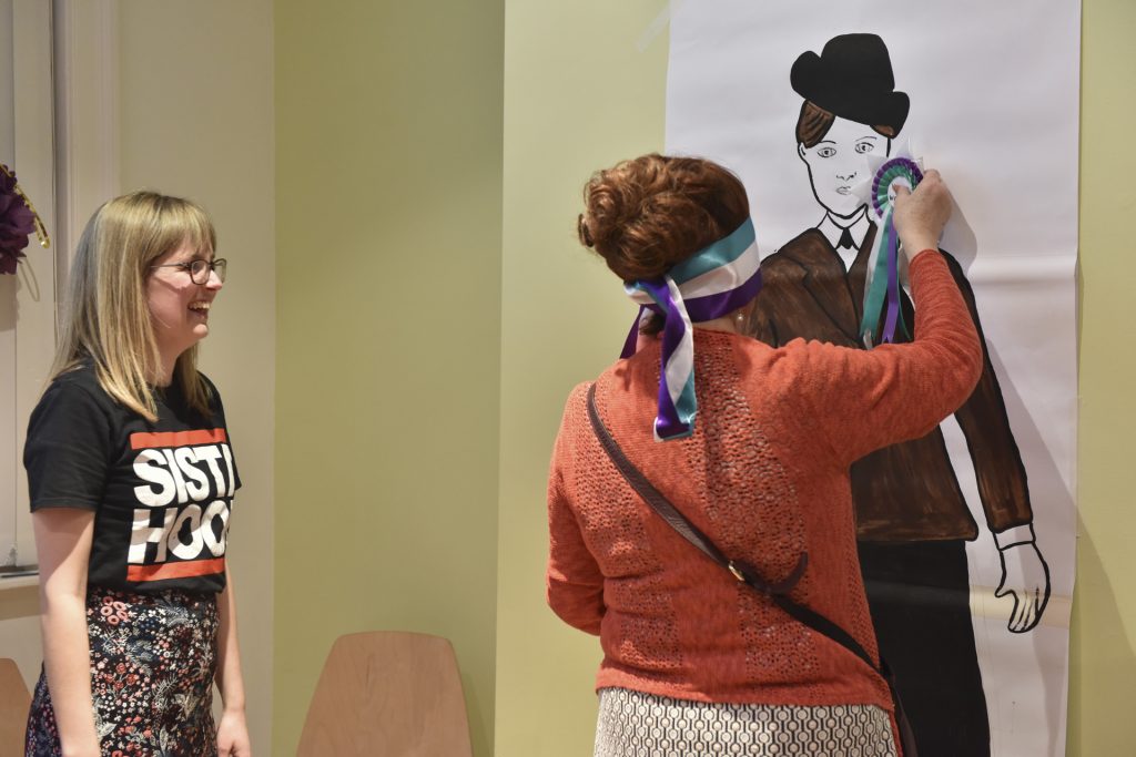 Two women are participating in a game. One is pinning a rosette on a drawing of a person wearing a hat, while the other watches and laughs. The setting appears to be a casual indoor environment.