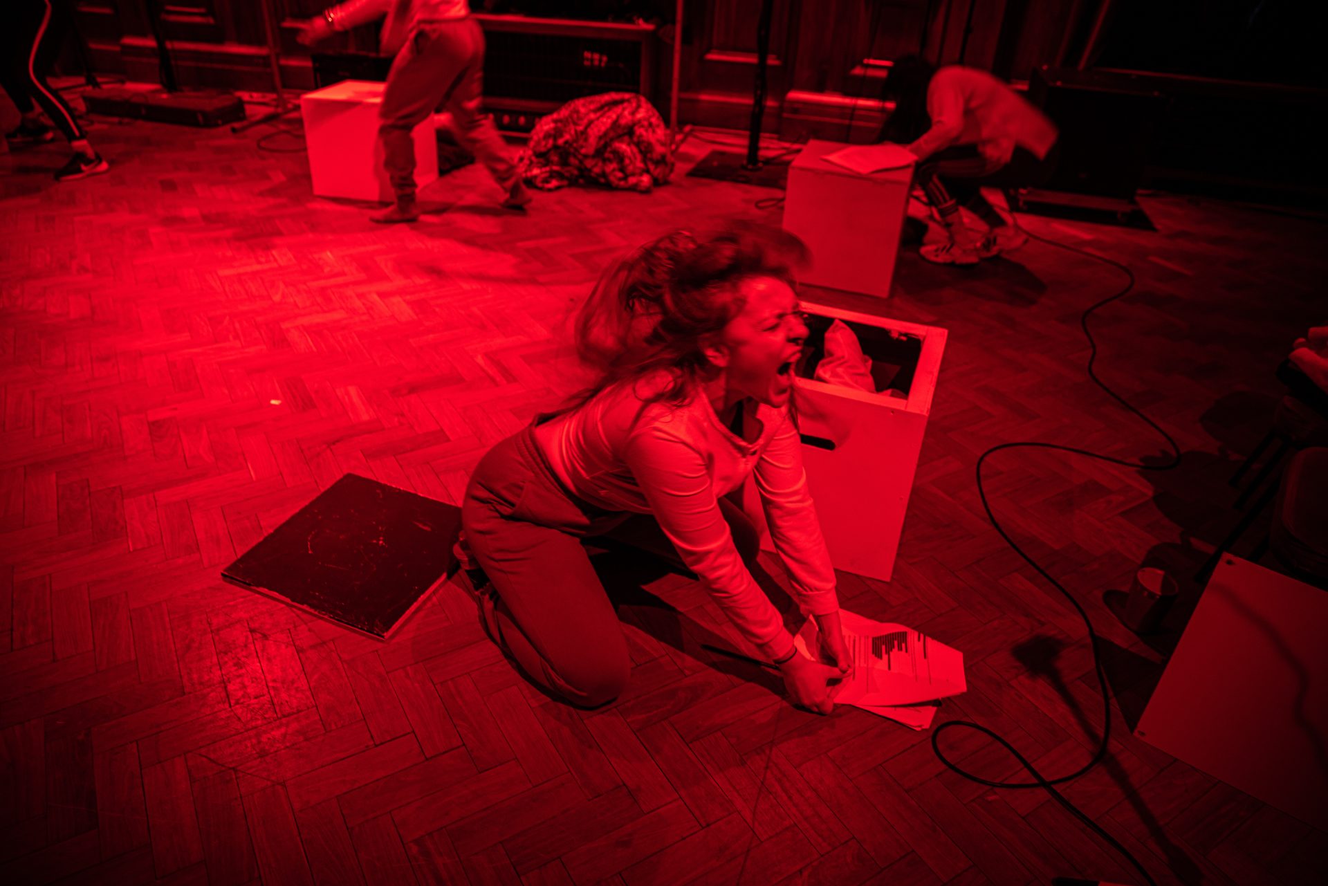 A person kneeling on a wooden floor under dramatic red lighting, appearing to be in an intense, emotional moment, holding sheets of paper. Other performers are visible in the background, each involved in separate actions with white boxes.