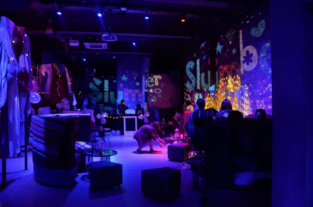 A dimly lit room features a cozy atmosphere with people sitting on couches and cushions under soft purple lighting. Walls are decorated with glowing words and twinkling fairy lights. A DJ booth is set up in the background.