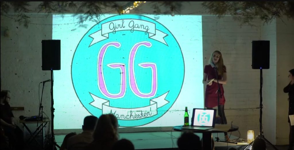 A woman stands on stage beside a large screen displaying the "Girl Gang Manchester" logo. The audience is seated in a dimly lit room, and there are small lights and a laptop on a table.