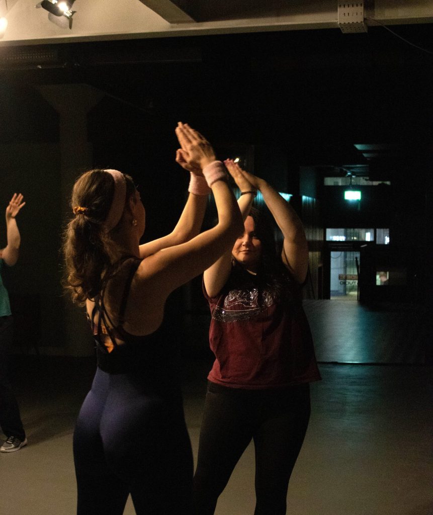 Two women clapping hands above their heads in a dimly lit room, appearing to enjoy a dance or exercise session. One wears a dark tank top and leggings, and the other a maroon T-shirt and black pants.