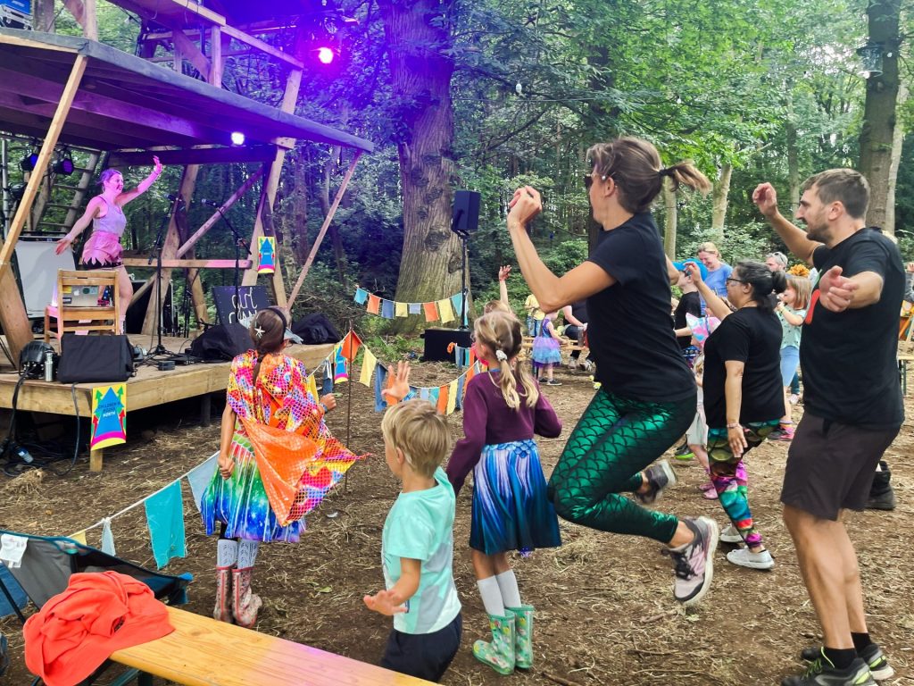 Adults and children dance energetically in front of an outdoor stage with a performer under colorful lights. The setting is a wooded area with vibrant decorations and festive flags. Everyone appears joyful, enjoying a lively, fun atmosphere.