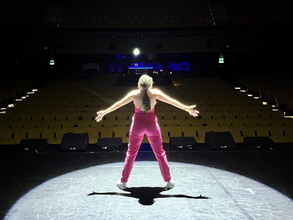 A person wearing a pink outfit stands in a spotlight on an empty stage, facing away from the camera, with arms outstretched. The theater seats are dimly lit in the background.