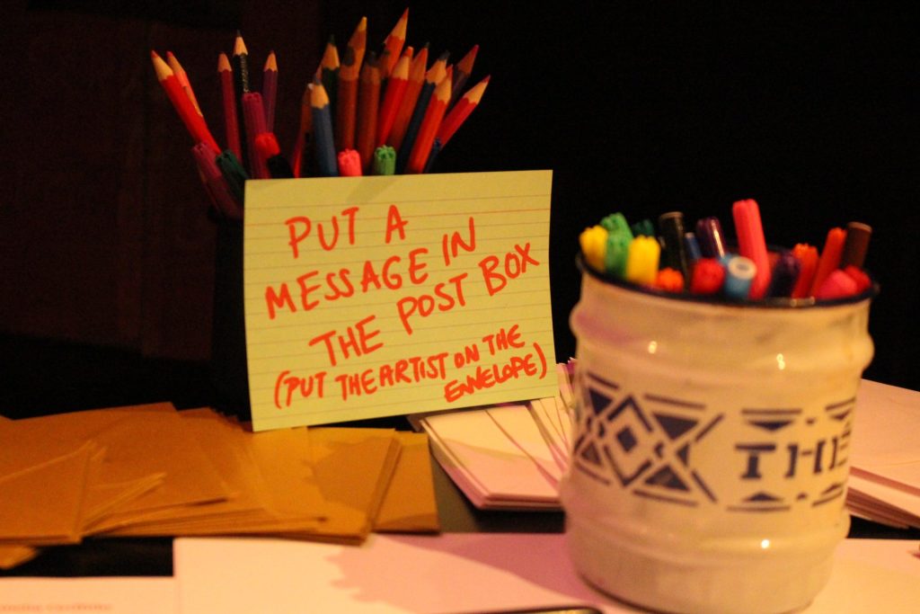 A table with colorful pencils in a black holder and multicolored pens in a white ceramic jar. A handwritten note on a green card reads, "Put a message in the post box (put the artist on the envelope)" next to some envelopes.