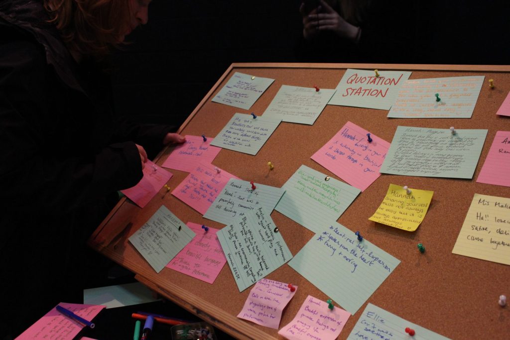 A person arranges colorful notes pinned to a corkboard labeled "Quotation Station." The notes contain handwritten messages and quotes. The scene is dimly lit, suggesting a thoughtful or creative atmosphere.