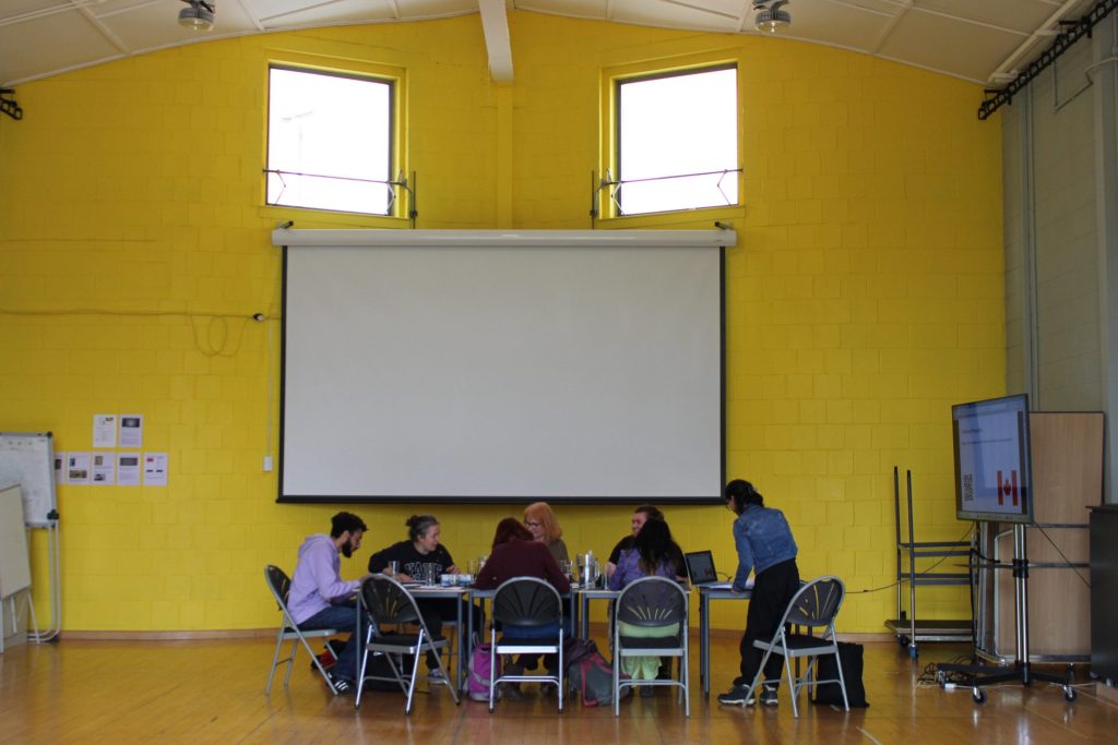 A group of people sit around a table in a room with bright yellow walls, facing a large screen. Two windows are above the screen, and there is a whiteboard on the left. A person stands near a monitor on the right.