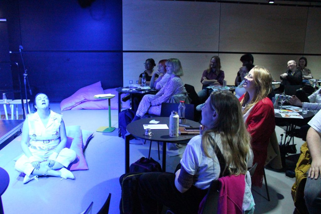 A group of people sitting at tables in a dimly lit room, watching a performer in white sitting cross-legged on stage, bathed in blue light. The audience appears engaged, some smiling, with notebooks and drinks on the tables.