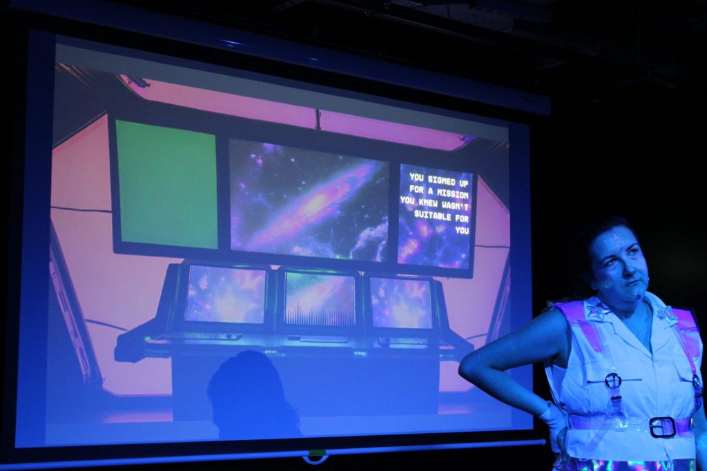 A person stands in front of a large screen displaying several monitors with space imagery and text reading, "You signed up for a mission you knew wasn't suitable for you." The room is dimly lit with a blue glow.